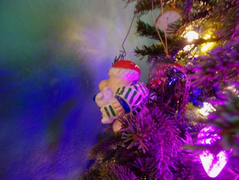 Small Christmas ornament of Ziggy, wearing a striped scarf and holding his dog, hanging from a tree and facing the wall.