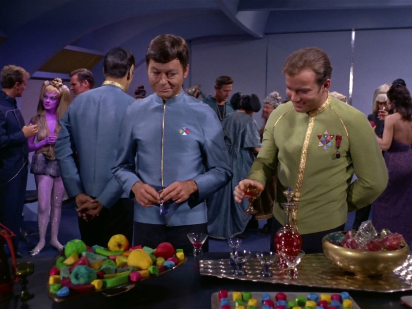 Cocktail party scene on the starship Enterprise. In the foreground Kirk and McCoy are joking around. In the background a purple woman in sparkling leggings and miniskirt, with a vest the color of her long hair so she doesn't quite look like she's wearing a top, smiles while looking at a guy with short, curly hair who's wearing a blue jumpsuit.