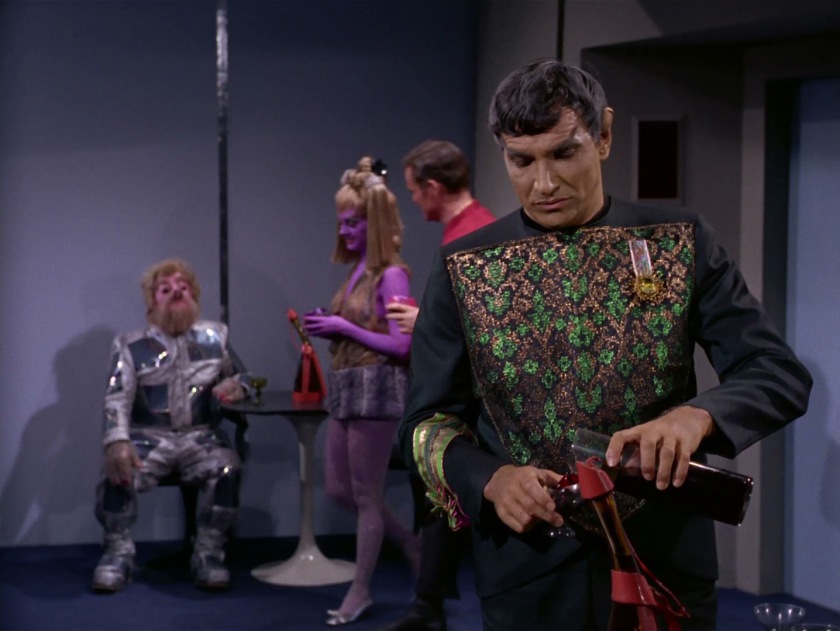 In the foreground Ambassador Sarek pours a drink from a very tall lab cylinder. In the background Ambassador Gav readies his courage to come up and yell at Sarek. In the mid-ground, a purple woman in sparkling tights and miniskirt walks across camera, accompanied by a short-haired man in red Star Fleet uniform.