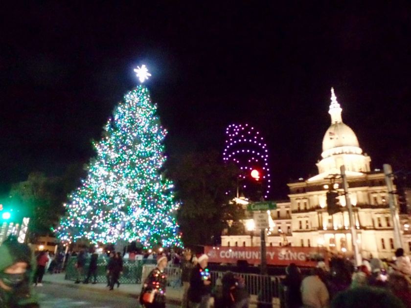 Photograph of the State Christmas Tree at its lighting, with the capitol in the background, and a fleet of drones in the distance arranged to make a picture of Santa Claus's face in the sky behind it all.