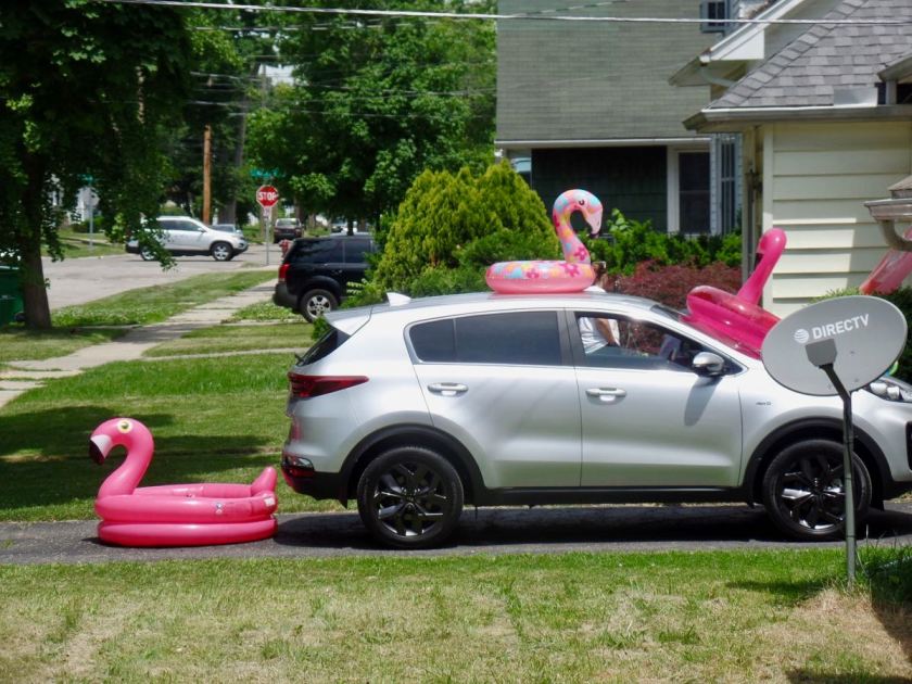 Photograph of a car parked in the driveway. Several inflatable flamingo pool rings are around: on the car's hood, on its windshield, on its roof, and a large one behind the car, another.