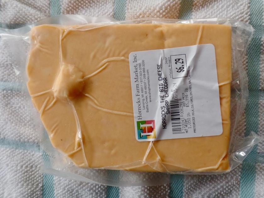 Photograph of a plastic-wrapped block of cheddar laid out on a tablecloth. It has a roughly rectangular shape, with the left side more of a curved arc, so that there's a faint resemblance to the outline of Montana if you're being generous. There is also a curious circular bulge in the center left, somewhere around where Missoula or Flathead Lake would be, that looks like a round security tag clinging to it.